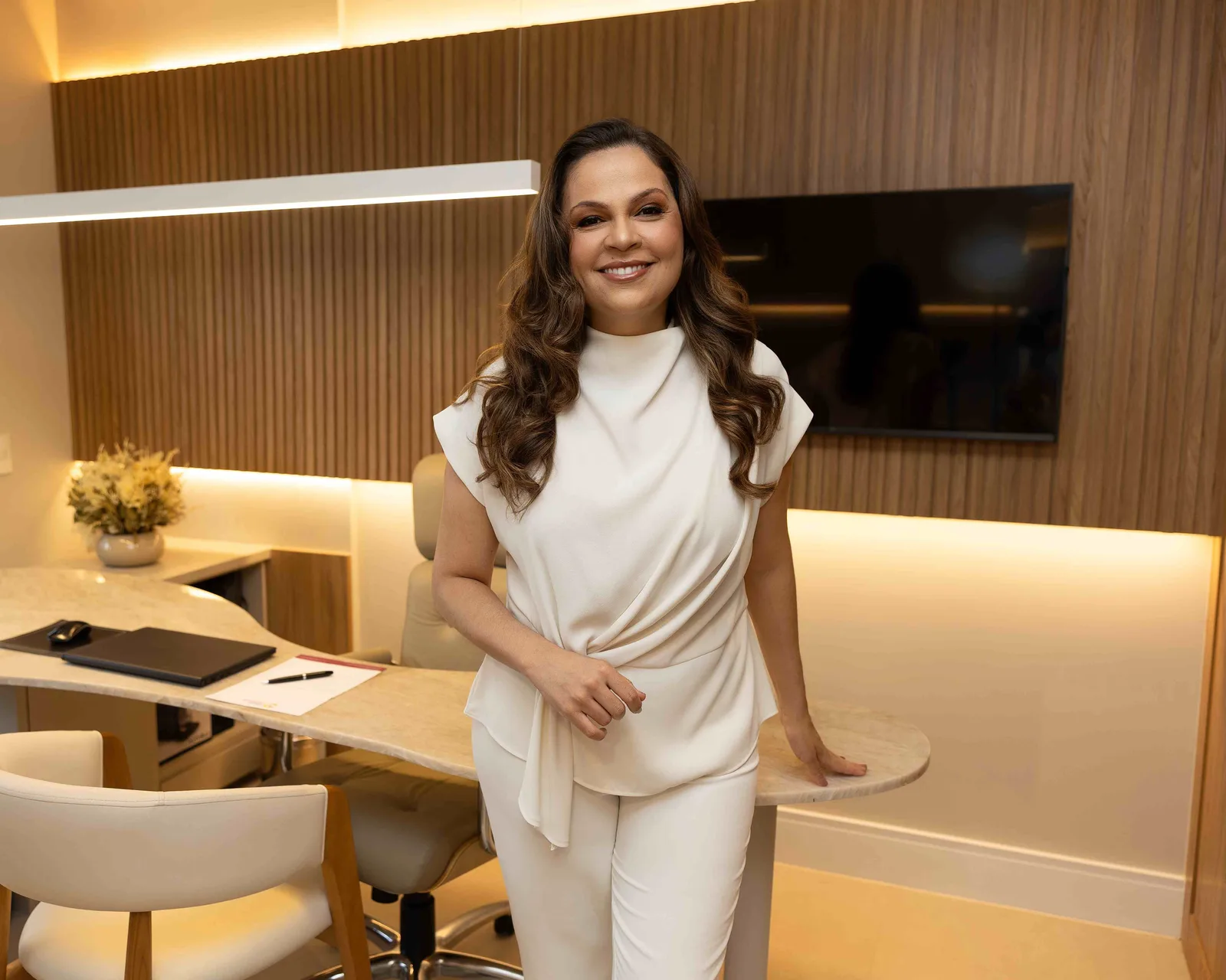 Dra. Gisane Cavalcanti, de pé em seu consultório, apoiada na mesa de atendimento